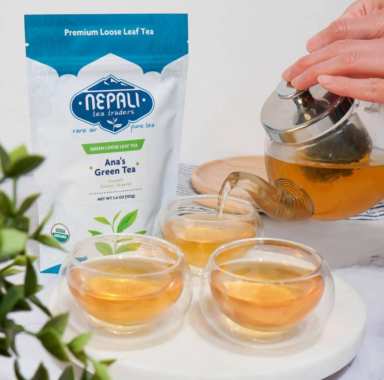 Pouring Ana’s Organic Green Tea from a glass teapot into double-wall cups beside a Nepali Tea Traders pouch—smooth, vegetal Nepal loose-leaf green tea.
