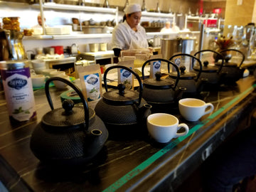 A row of black teapots and white cups set up for tea service with Nepali tea products in a professional kitchen.