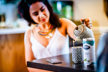 A woman prepares a soothing cup of tea from a Nepali Tea Traders canister. She pours the white tea from a teapot, illustrating a calming evening ritual to promote sleep.