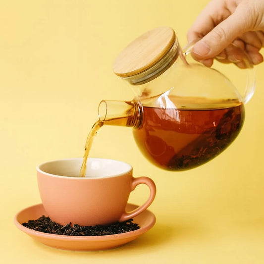 Black loose leaf tea being poured from a glass teapot into a coral-pink teacup with matching saucer, on a bright yellow background