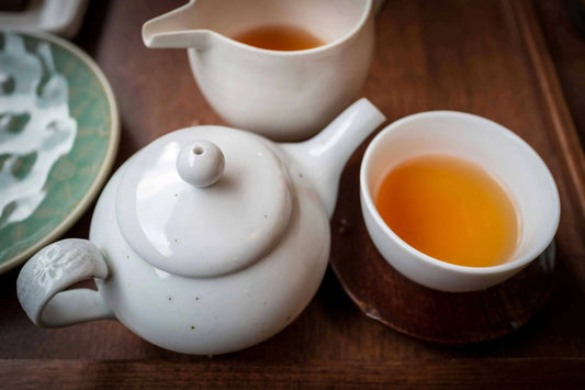 A close-up of a traditional white ceramic teapot and cup filled with golden loose leaf tea, set on a wooden tea tray beside a green patterned dish.