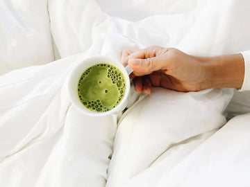 Hand holding a mug of frothy matcha green tea latte while relaxing in bed