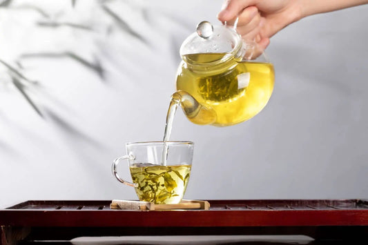 A hand pouring freshly brewed loose-leaf green tea from a clear glass teapot into a glass cup on a wooden tea tray, featuring a minimalist aesthetic.