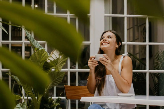 A smiling woman sitting at a white cafe table, holding a cup of Nepali loose leaf tea and looking upward, framed by lush green indoor plants.