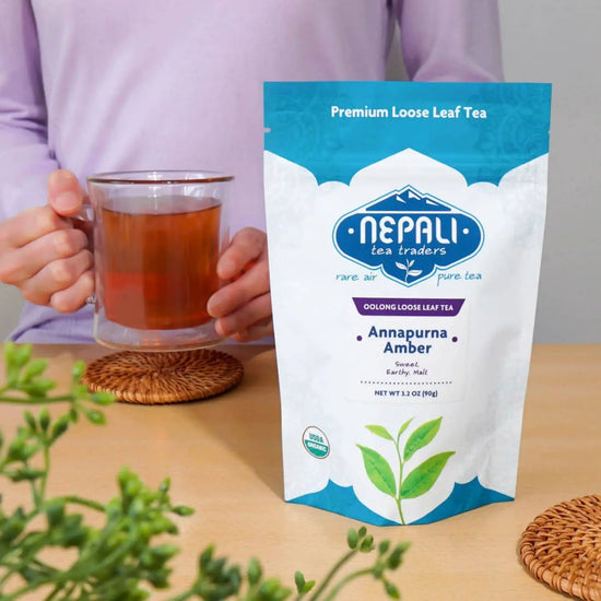 Annapurna Amber Organic Oolong Tea by Nepali Tea Traders—USDA Organic pouch on a table with a person holding a glass mug of brewed oolong.