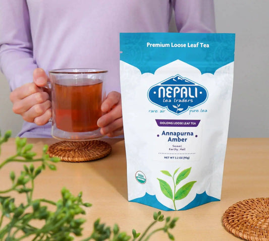 Annapurna Amber Organic Oolong Tea by Nepali Tea Traders—USDA Organic pouch on a table with a person holding a glass mug of brewed oolong.