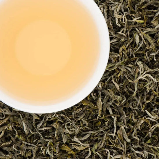 A white cup of brewed Jade Spring white tea with a pale gold liquor, surrounded by premium silver-green loose tea leaves from Nepal."