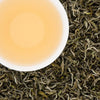 A white cup of brewed Jade Spring white tea with a pale gold liquor, surrounded by premium silver-green loose tea leaves from Nepal."