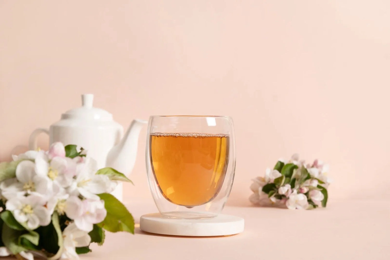 Aromatic blended Nepali tea in a double walled glass cup with flowers and a white teapot on a blush background