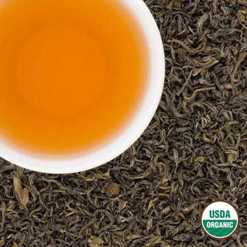 Amber liquor of Pokhara Classic organic green tea in a white cup over loose leaves, USDA Organic seal