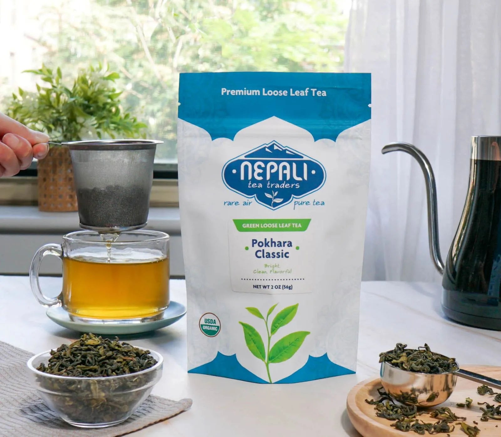 Pokhara Classic Organic Green Tea from Nepal—Nepali Tea Traders pouch with brewed green tea, loose leaves, and a strainer over a glass mug.