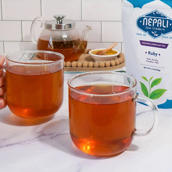 Ruby Organic Oolong Tea from Nepali Tea Traders—two clear mugs of brewed oolong beside the product pouch, with a glass teapot in the background.