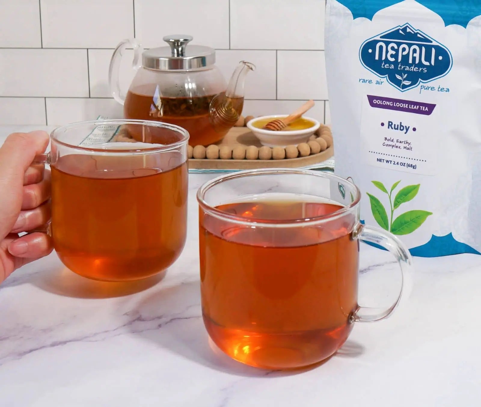 Ruby Organic Oolong Tea from Nepali Tea Traders—two clear mugs of brewed oolong beside the product pouch, with a glass teapot in the background.