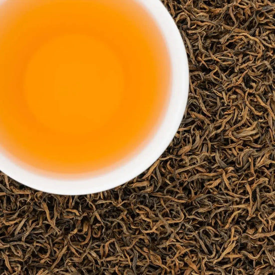 A white cup of brewed Sagarmatha Gold black tea showing a vibrant honey-colored liquor next to dry loose leaves.