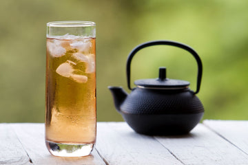 Tall glass of iced tea with ice cubes beside a black teapot on a wooden table